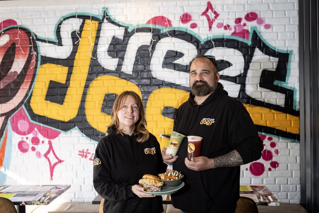 Two people standing in front of graffiti saying Street Dogs, holding burgers, chips and drinks.
