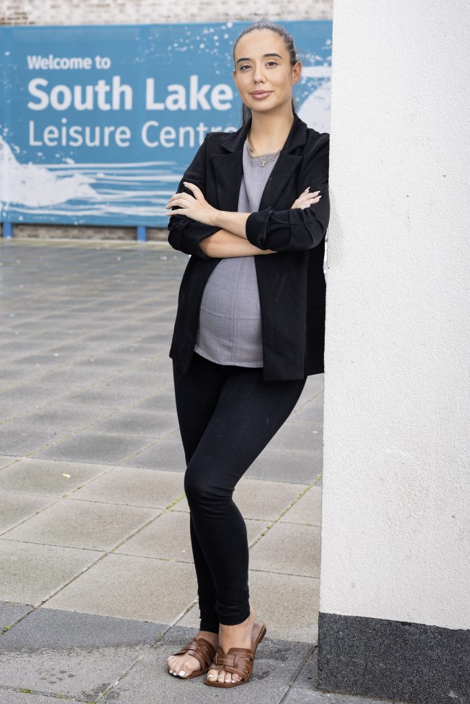 A woman standing, leaning against a wall with a sign in the background that reads 'South Lake Leisure Centre'.